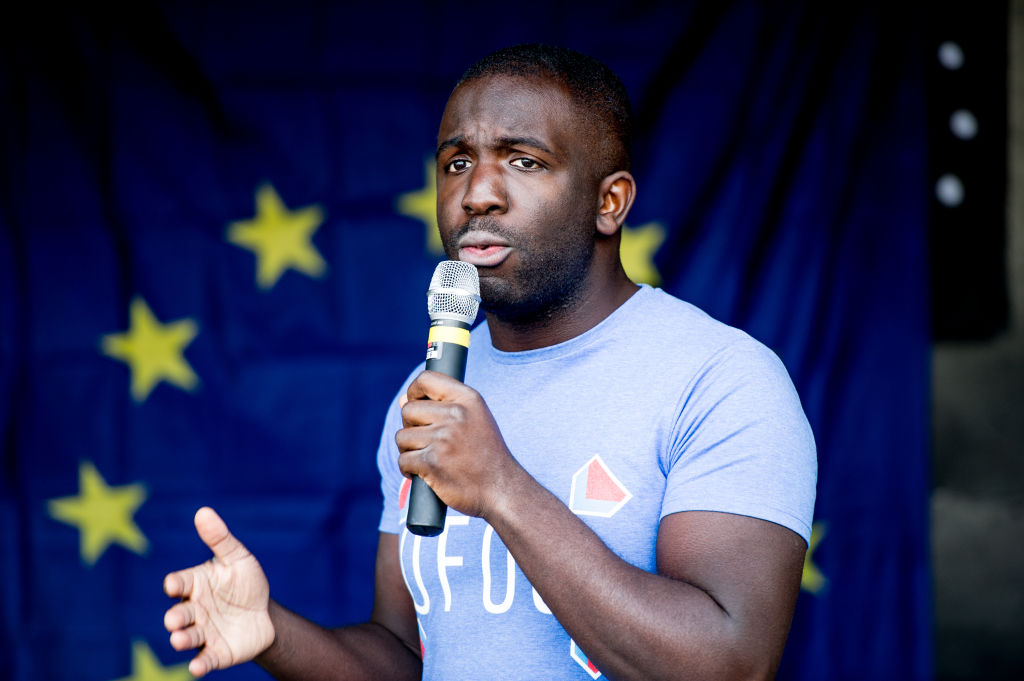 Femi Oluwole speaks into a microphone at an anti-Brexit march. Behind him is the blue and yellow EU flag