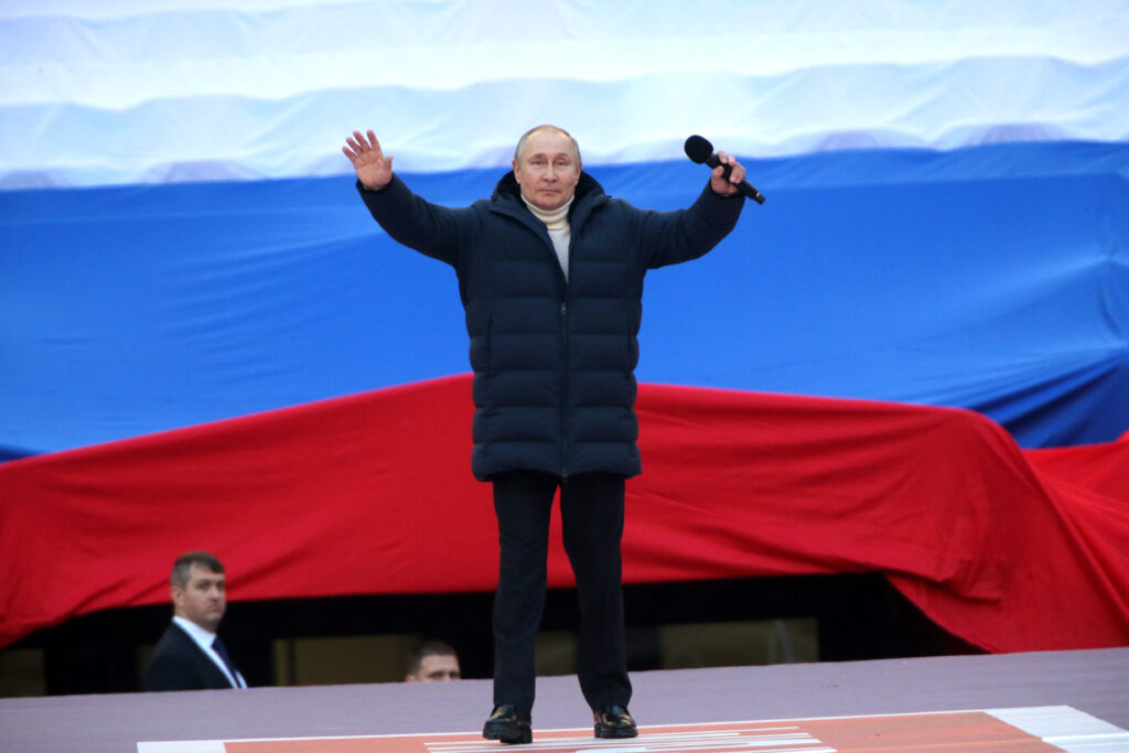 President Putin stands on stage in front of the colours of the Russian flag with his arms in the air at a concert to mark the Russian annexation of Crimea