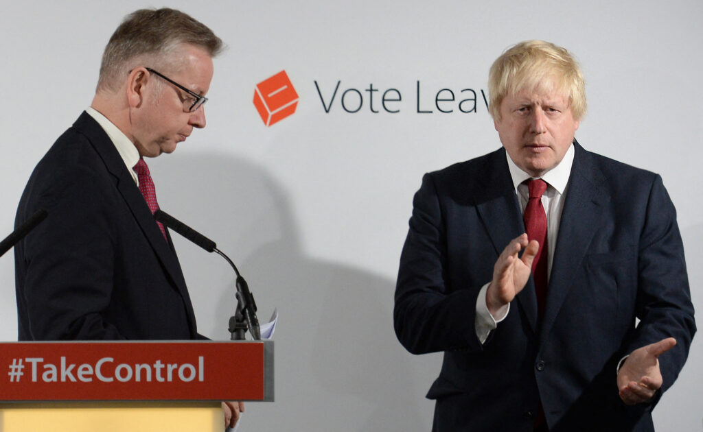 Michael Gove stands at a podium with the words TakeControl written on it. Boris Johnson stands to one side clapping. In the background is the Vote Leave logo.