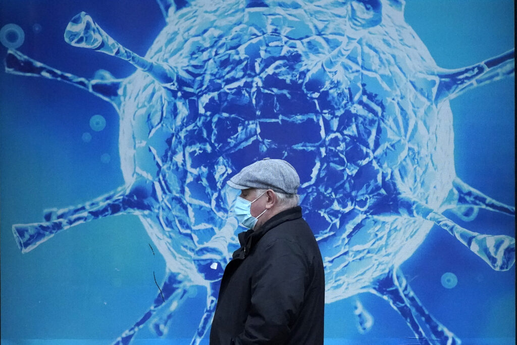 A man wearing a facemask walks in front of an image of the Covid-19 virus